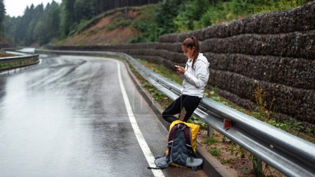 Photo for Young caucasian woman with backpack standing on wet road among mountains and using smartphone. Female hiker searching for route on cell phone. - Royalty Free Image