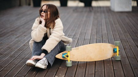 Téléchargez les photos : Femme hipster caucasienne en lunettes de soleil, manteau de fourrure et jean foncé assis sur le toit près de son skateboard. passe-temps extrême préféré de la jeune brune. - en image libre de droit