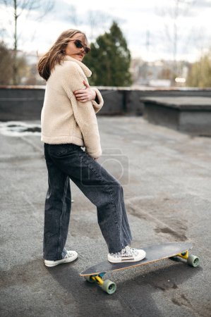 Photo for Confident good looking woman with short brown hair standing on skateboard on roof top. Young female hipster enjoying favorite extreme hobby outdoors. - Royalty Free Image