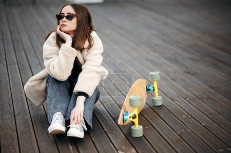 Téléchargez les photos : Femme hipster caucasienne en lunettes de soleil, manteau de fourrure et jean foncé assis sur le toit près de son skateboard. passe-temps extrême préféré de la jeune brune. - en image libre de droit
