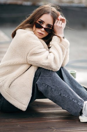 Téléchargez les photos : Femme caucasienne à la mode assise sur le sol en bois de roop top et regardant avec confiance la caméra à travers des lunettes de soleil à la mode. Concept de jeunesse, de personnes et de modes de vie. - en image libre de droit