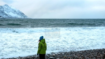 Photo for Lofoten islands, Senja region, Norway. Winter. Young girl in green jacket and blue hat stands with her back to ocean. Small wave. In background. snowy mountains. Concept of tourism. - Royalty Free Image