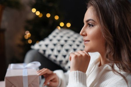 Téléchargez les photos : Portrait de la jeune femme posant avec des cadeaux le matin après Noël. Femme est assis dans le salon à l'arrière-plan de l'arbre de Noël - en image libre de droit