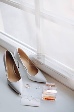 Foto de Vista de cerca de los detalles de la boda. Zapatos de la elegancia de la novia, anillos, perfume y la invasión layflat en alféizar de la ventana blanca - Imagen libre de derechos