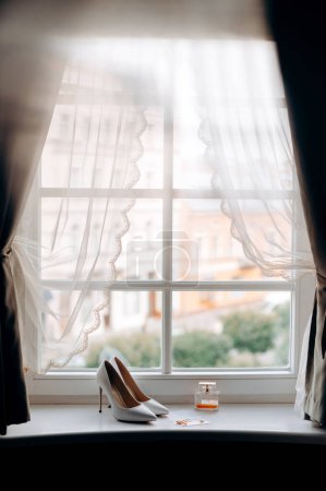 Foto de Anillos de boda, zapatos y parfums en el alféizar de la ventana de la habitación de hotel brillante - Imagen libre de derechos