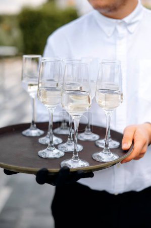 Téléchargez les photos : Serveur homme en chemise blanche tenant plateau avec verres de vin blanc ou champagne sur fond de terrasse - en image libre de droit