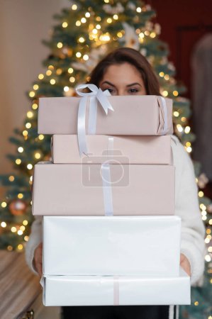 Foto de Vista vertical de la joven feliz sosteniendo una gran cantidad de regalos cerca del árbol de Navidad en casa. Concepto de vacaciones y tradiciones - Imagen libre de derechos