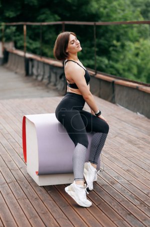 Photo for European sportswoman sitting and relax on fitness mat on white cube. Wooden terrace or balcony. Young beautiful girl with closed eyes wear sportswear and sneakers. Concept of sport at outdoors - Royalty Free Image