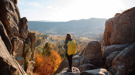 Photo for Outdoor recreation. Young girl in sports clothes, in gray jacket and backpack on her shoulders stands back, on stone mountain, in background small village in mountains. Autumn. Concept of tourism. - Royalty Free Image