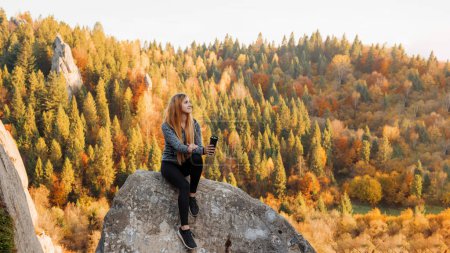Photo for Outdoor recreation. Young girl in sports clothes, in gray jacket, with thermos in her hands, sitting on stone mountain, looking away, in background autumn forest and mountains. Copy space - Royalty Free Image