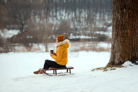 Foto de Mujer joven con chaqueta de invierno amarilla sentada en un trineo y bebiendo té de la taza termo en el parque de invierno. - Imagen libre de derechos