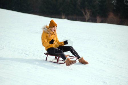 Foto de Mujer joven sonriente con chaqueta de invierno amarilla cabalgando colina abajo en el trineo en el parque de invierno nevado. Chica trineo abajo - Imagen libre de derechos