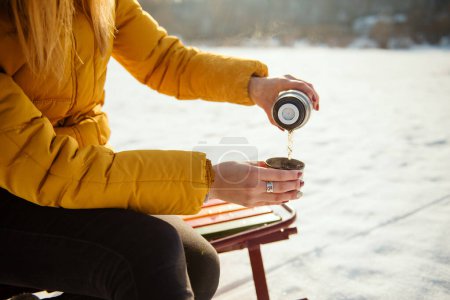 Téléchargez les photos : Femme en veste d'hiver jaune assise sur le traîneau et buvant du thé chaud du thermos. Fille versant tasse avec thé chaud - en image libre de droit