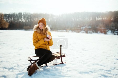 Foto de Mujer sensual joven en chaqueta de invierno amarilla sentada en el trineo y bebiendo té caliente del termo en el parque nevado - Imagen libre de derechos