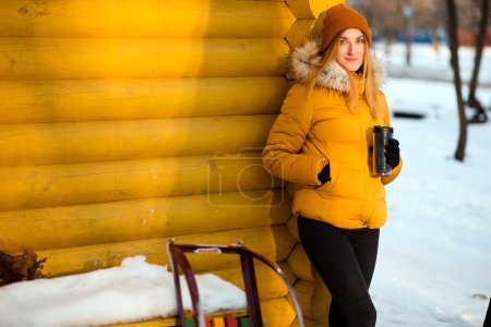 Téléchargez les photos : Belle jeune femme en veste d'hiver avec thermotasse et traîneau debout près du mur en bois jaune à l'extérieur dans le parc enneigé - en image libre de droit