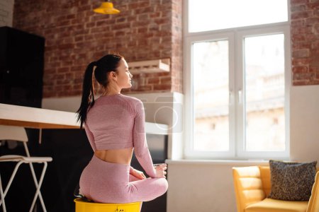 Photo for Side view of charming slender woman in sport clothes resting on yellow barrel and holding glass of fresh water. Recreation during domestic workout. - Royalty Free Image
