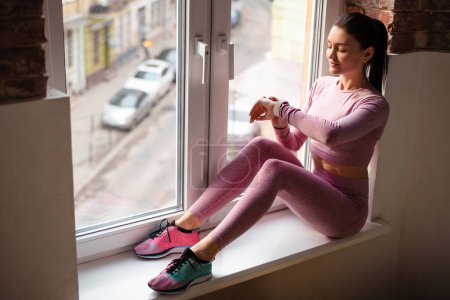 Téléchargez les photos : Une femme sportive en tenue de sport est assise près de la fenêtre et utilise une montre intelligente à la maison dans le salon. Concept de sport et de loisirs. - en image libre de droit