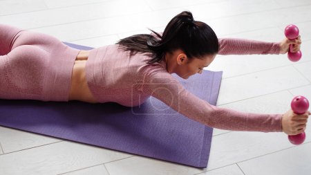 Foto de Mujer joven deportiva en ropa deportiva rosa acostada en una esterilla de yoga y brazos de entrenamiento con mancuernas. Caucásico morena teniendo doméstica entrenamiento durante el tiempo libre. - Imagen libre de derechos