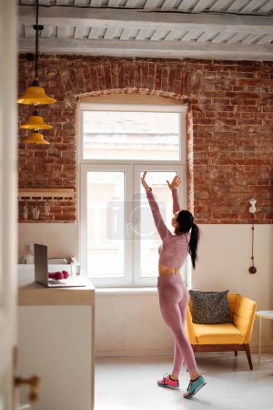 Photo for Back view of fitness young woman standing near window at modern apartment with outstretched arms. Pretty caucasian lady relaxing during domestic workout. - Royalty Free Image