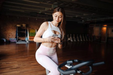 Photo for Portrait of smiling fitness girl using mobile phone after training on exercise bike in evening at modern gym. Portrait of model, fit sporty woman, at gym engaged in a stationary bike - Royalty Free Image