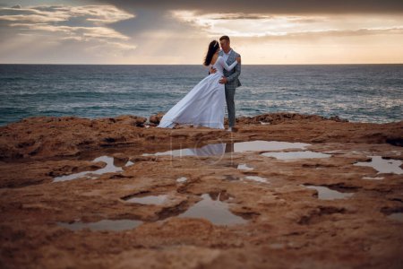 Foto de Los recién casados posan en ropa de boda en las rocas junto al mar. Hermosa joven pareja tiernamente abrazando. Boda de recién casados en la isla
. - Imagen libre de derechos