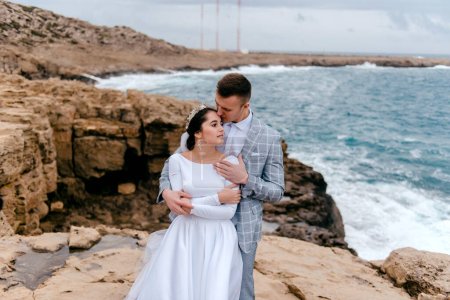 Foto de Retrato de recién casados felices en la costa rocosa del mar. El novio besa tiernamente a la novia. Feliz día de la boda en Chipre
. - Imagen libre de derechos