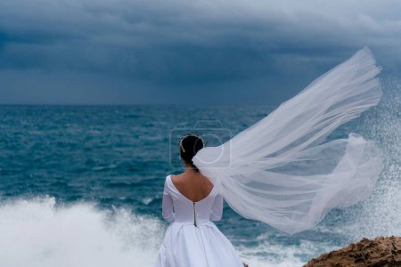 Téléchargez les photos : Vue arrière de la mariée brune en robe de mariée blanche et voile nuageux par une journée nuageuse. Romantique belle mariée posant près de la mer avec des vagues. Le voile nuptial vole dans le vent
. - en image libre de droit