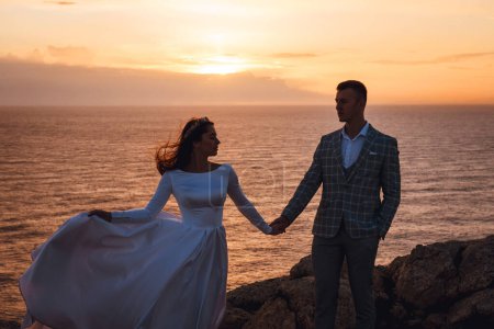 Téléchargez les photos : Les mariés marchant le long de la côte rocheuse au coucher du soleil. Silhouette de jeunes mariés amoureux sur fond de mer. Mariage pour deux à Chypre
. - en image libre de droit