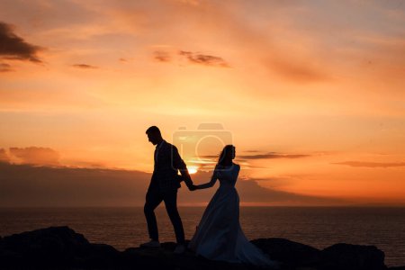 Foto de Retrato de una hermosa novia y novio al atardecer en Chipre. Pareja joven sobre un fondo de mar y cielo naranja. Boda y amor concepto
. - Imagen libre de derechos