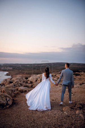 Foto de Vista posterior de la joven pareja de boda, novia y novio, caminando en una playa rocosa en la noche Chipre. Hermoso día de la boda, tiempo para dos
. - Imagen libre de derechos