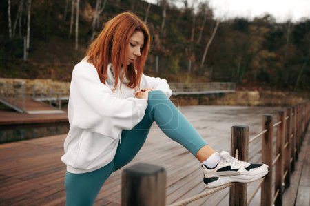 Foto de Mujer joven en forma y deportiva con el pelo rojo en ropa deportiva estirando sus piernas después del entrenamiento en el muelle de madera en el lago cerca del bosque verde. - Imagen libre de derechos