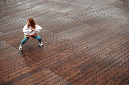 Téléchargez les photos : Jeune femme en forme sportive aux cheveux roux dans les squats de vêtements de sport avec bande élastique de remise en forme sur jetée en bois. Concept sportif, mode de vie sain - en image libre de droit