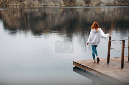 Téléchargez les photos : Femme sportive avec les cheveux roux en sweat à capuche blanc et leggings bleus debout à quai en bois près du lac et bénéficiant d'une vue paisible. Femme en forme tenant le téléphone et la bouteille. Vue arrière - en image libre de droit