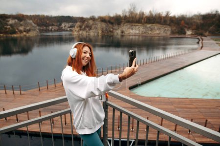 Téléchargez les photos : Joyeux sourire rousse fille aux cheveux avec une bouteille d'eau et écouteurs sur sa tête prend un selfie. Carrière de Zakrzwek, Cracovie. Piscine flottante. Jour d'automne nuageux - en image libre de droit