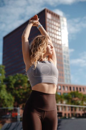 Photo for Young sporty female workout before fitness training session warming up and stretching her arms over street background - Royalty Free Image