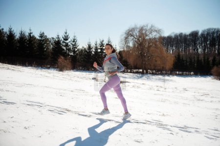 Photo for Side view of young satisfied motivated and focused sporty active girl with a fluffy ponytail in winter sportswear running in the snowy nature - Royalty Free Image