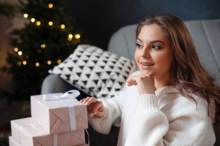 Téléchargez les photos : Portrait de la jeune femme posant avec des cadeaux le matin après Noël. Femme est assis dans le salon à l'arrière-plan de l'arbre de Noël - en image libre de droit