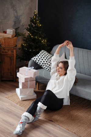 Téléchargez les photos : Vue verticale de la femme positive étirant les mains le matin après Noël. Femme est assis dans le salon à l'arrière-plan de l'arbre de Noël - en image libre de droit