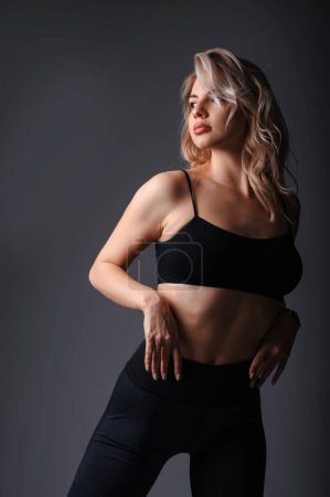 Photo for Portrait of a beautiful young blonde woman in black top and leggings. Girl in sportswear posing on black studio background - Royalty Free Image