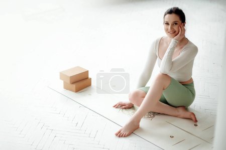 Téléchargez les photos : Portrait de belle jeune femme en T-shirt blanc et leggings verts assis sur le tapis de yoga et souriant à la caméra. Cubes pour des exercices de streching près d'elle - en image libre de droit