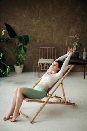 Téléchargez les photos : Belle jeune femme assise sur une chaise Sunbad dans un salon confortable et relaxant après l'entraînement ou le yoga. Concept sain et sportif - en image libre de droit