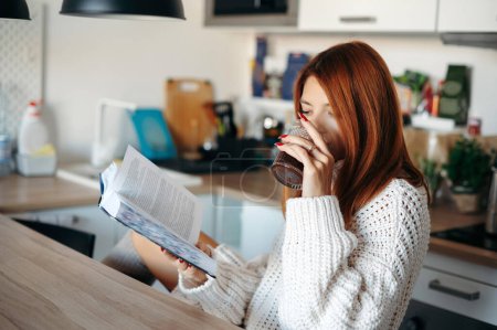Foto de Joven mujer pelirroja en ropa acogedora y cálida bebiendo café y leyendo libro en la cocina en casa - Imagen libre de derechos