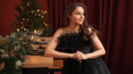 Foto de Mujer joven en vestido negro posando con la decoración del árbol de Navidad en casa y sonriendo. Luces y bokeh. Concepto de invierno y vacaciones - Imagen libre de derechos
