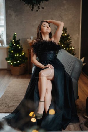 Foto de Hermosa chica en vestido de noche sentada en el sillón cerca del árbol de Navidad. Fondo interior de lujo. Vacaciones de Navidad, celebración, concepto de regalos - Imagen libre de derechos