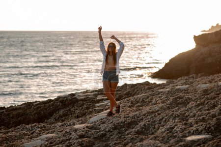 Foto de Mujer viajera atractiva en camisa blanca y pantalones cortos de pie sobre una roca junto al mar y levantó las manos. Concepto de viaje y aventura - Imagen libre de derechos