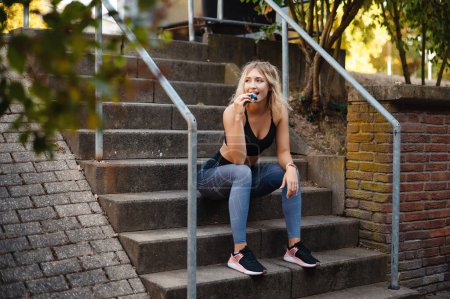 Photo for Smilling fitness woman in sportswear eating fitness bar while sitting on stairs outdoors. Blonde sporty girl resting after workout. Healthy and sport concept - Royalty Free Image