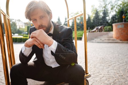 Photo for Confident bearded groom in stylish black suit sitting in golden luggage trolley outdoors. Warm summer weather outdoors. Wedding day. - Royalty Free Image