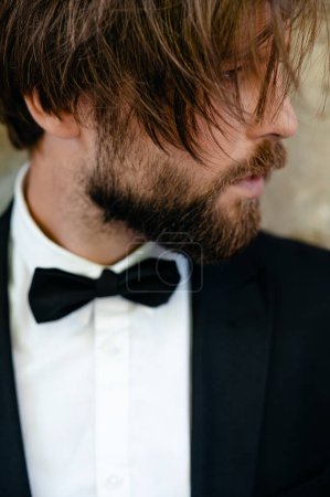 Foto de Elegante novio caucásico en traje de moda con pajarita negra posando al aire libre. Retrato del guapo barbudo. Día de la boda. - Imagen libre de derechos