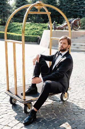 Photo for Stylish handsome groom wearing elegant suit sitting in hotel luggage trolley outdoors. Bearded caucasian man posing on camera. - Royalty Free Image
