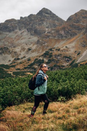 Foto de Elegante mujer excursionista con gafas de sol y mochila caminando en las montañas Hight Tatras, Polonia. Magestic gran roca en el fondo - Imagen libre de derechos